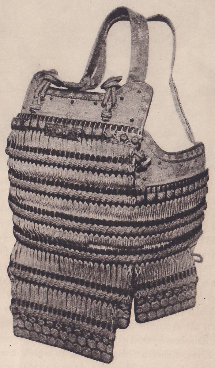 Black-and-white photograph of a laced body piece, set on a pale ground. A flat plate across its top has knotted cords at both ends, and two long straps rise from it, one curving over to the left and one hanging at the right. Below the plate come many horizontal bands of closely set pale-tipped lacing, divided by twisted cords, and the lower edge splits into two flaps, each ending in a row of round shapes.