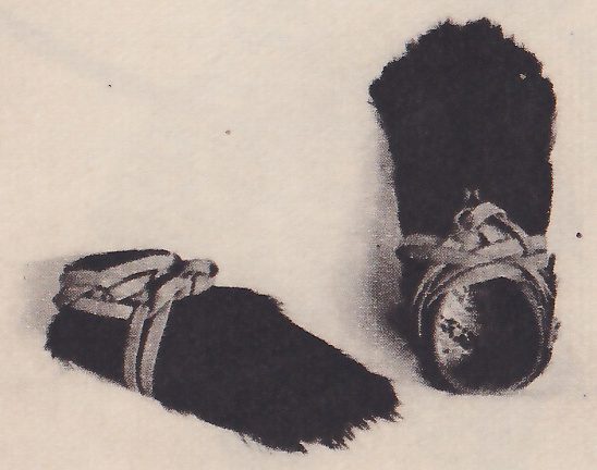 Black-and-white photograph of two shaggy dark objects on a pale ground. One lies at the lower left with pale straps wound across its top. The other stands tilted on end at the right, with a pale cord wound around its middle and a paler round patch at its lower front.