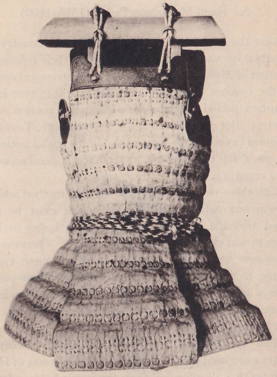 Black-and-white photograph of a body piece hung from a flat pale board by two knotted cords at the top. Below a dark curved panel, a tall body of pale horizontal bands with rows of dark round dots narrows toward a braided cord at the waist, and a wide skirt of similar bands flares out below, with a dark side at the right.