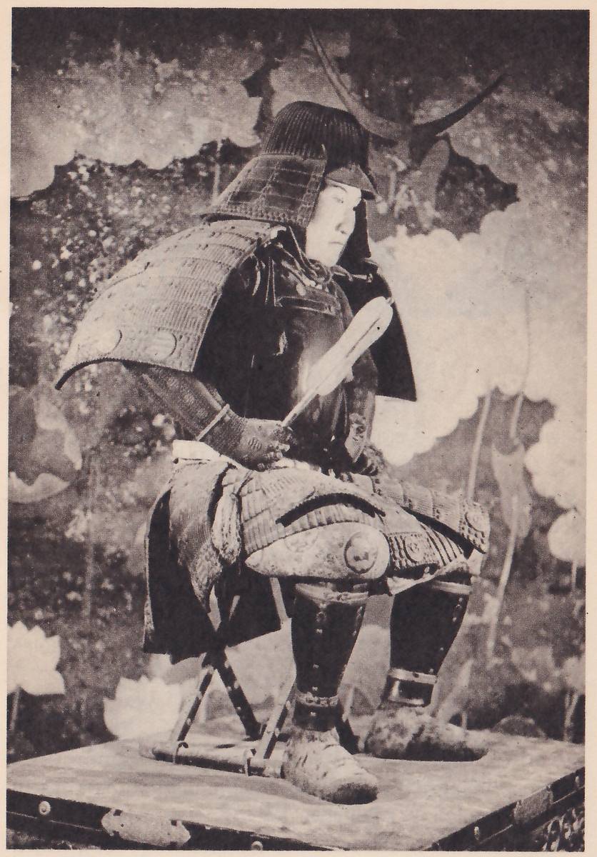 Black-and-white photograph of a figure in armour sitting on a seat with crossed legs on a low platform with metal edge fittings, in front of a backdrop of large pale flowers and leaf shapes. A dark helmet with a curved crest rising behind it sits above a pale face turned toward the right. A broad flat shoulder piece spreads out at the left, and a long pale flat object slants across the chest toward a hand at the left. Banded skirt pieces cover the lap, with dark leg pieces and rounded shoes below.