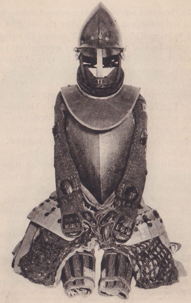 Black-and-white photograph of a suit of armour set up in the shape of a figure, with two hand coverings resting at the front. A pointed dome helmet with a rim sits at the top over a dark face covering with a pale cross-shaped gap. A broad pale collar spreads over a smooth curved body plate with a ridge down its middle. Sleeves of fine mesh hang down both sides, mesh covers the thighs, and vertically ridged leg pieces show at the bottom.
