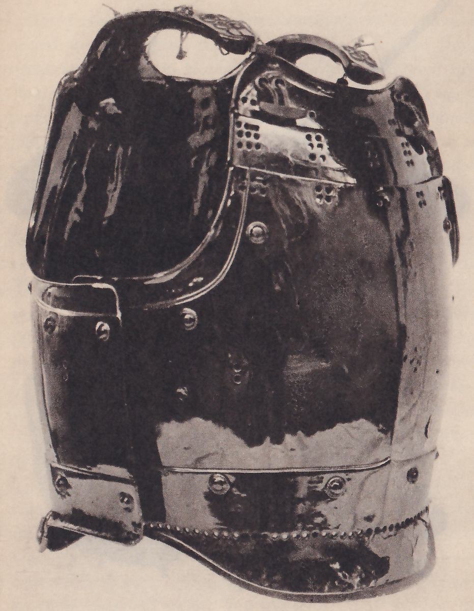 Black-and-white photograph of a glossy dark body piece with a smooth rounded front, seen at an angle. Two large loops rise at its top, the one at the left larger and open. Rows of round studs run across its middle and along its lower edge, small clusters of holes show on patches near the top, and a flat plate sticks out at the lower left.
