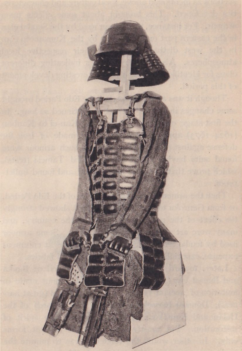 Black-and-white photograph of a suit of armour on a stand. A dark helmet with a flared lower edge sits at the top on a pale cross-shaped post. Below it a body piece of rows of dark and pale oblong panels, bound by cords, has sleeves of fine mesh hanging at both sides, with hands resting at the front and a skirt of fine mesh at the bottom. A pale rectangle shows behind at the right.
