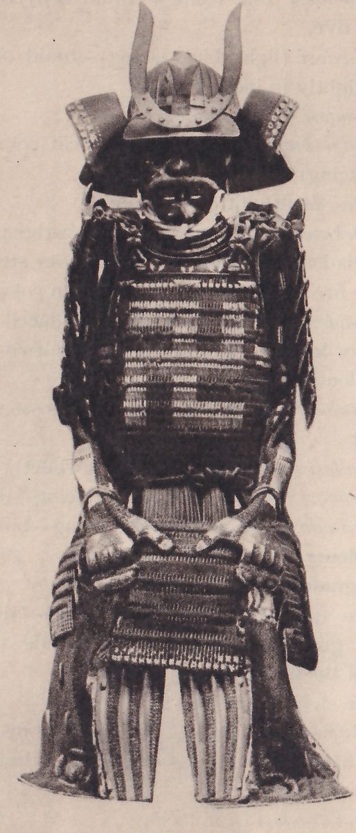 Black-and-white photograph of a standing suit of armour. A dark helmet with two curved prongs rising from its front has broad flared flaps at both sides, over a dark face covering with a pale cord below it. The body is horizontal bands of pale and dark lacing with a stepped pale pattern across the chest. Two hand coverings with pale fingers rest at the front of the waist, and vertically striped leg pieces show at the bottom between dark side pieces.