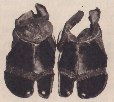 Black-and-white photograph of two dark flat pieces side by side. Each has a curled flap rising from one upper corner, a pale stitched border around a darker central panel, and a split at the bottom into two narrow tips, each with a small pale round dot. The left one has a round disc at its upper left.
