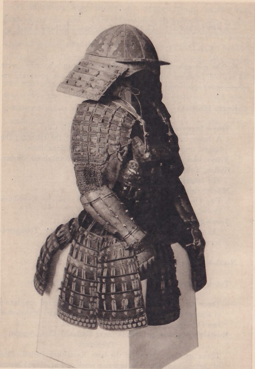 Black-and-white photograph of a suit of armour on a pale stand. A ribbed dome helmet with a flat brim at the right and a deep guard of laced bands at its left rises above a broad shoulder piece of small rectangular plates. A sleeve covered in fine mesh runs down to a banded forearm piece, and a skirt of laced bands with a studded lower edge hangs below, with a dark mass behind at the right.