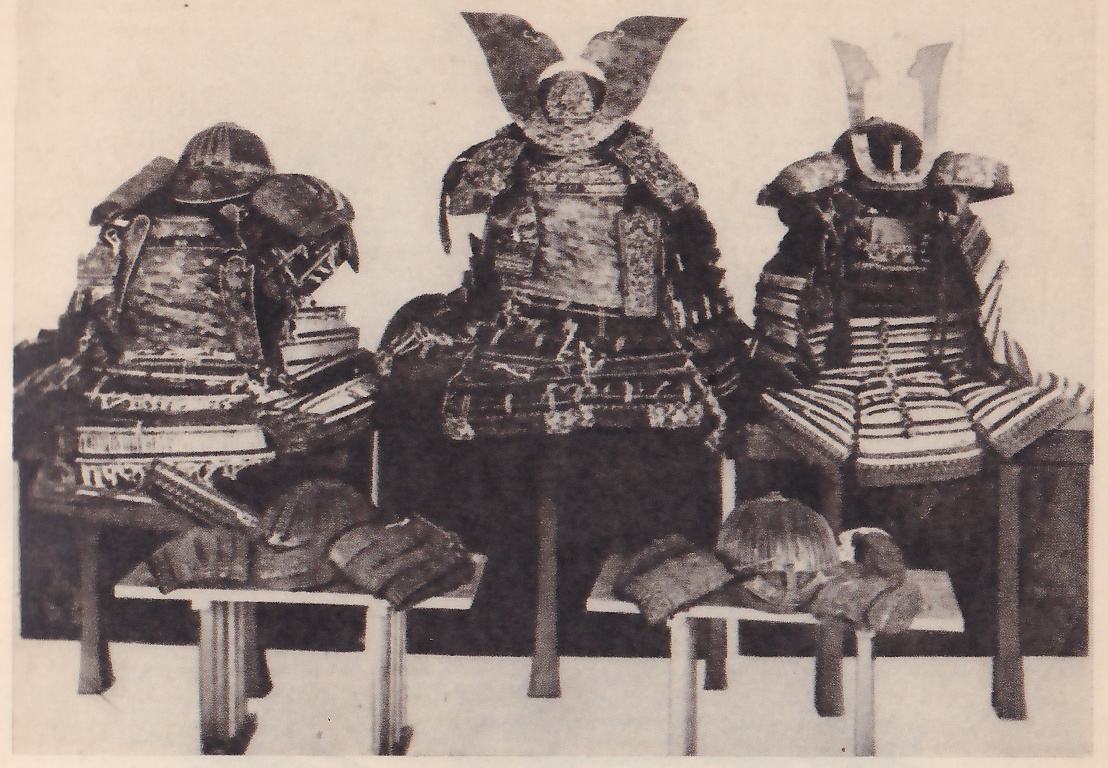 Black-and-white photograph of three suits of armour on stands in a row, with two low tables in front holding smaller pieces. The one at the left has a ribbed helmet between broad shoulder pieces. The middle one has two broad curved plates rising at the top above patterned shoulder pieces and a dark skirt of laced bands. The one at the right has two thin pale prongs rising from its helmet and wide skirt pieces of pale and dark stripes.