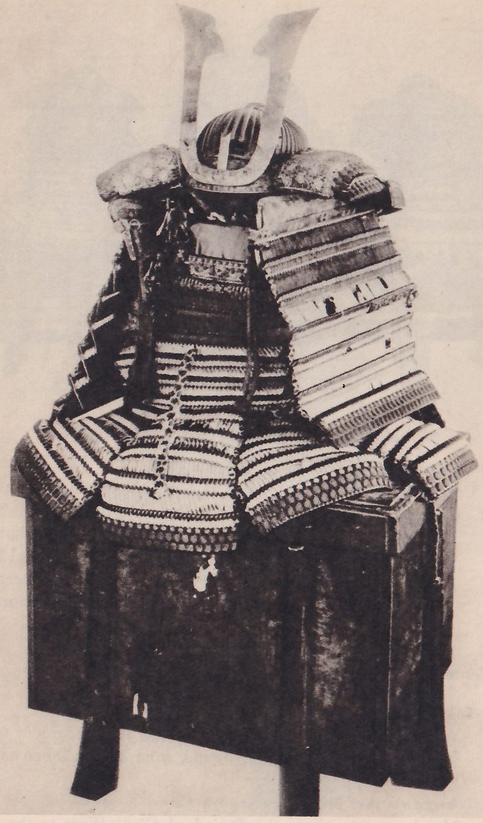 Black-and-white photograph of a suit of armour on a tall dark box, with two pale prongs rising in a V from the helmet at the top. A broad flat shoulder piece of pale bands stands at the right, and a banded skirt of pale and dark rows with a lower edge of small hexagons spreads over the top of the box. A small pale shape shows below the skirt.