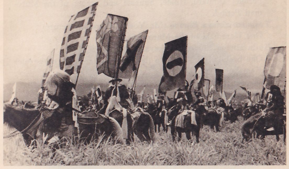 Black-and-white photograph of a crowd of figures on horseback in a grassy field, with many tall banners on poles held up above them. At the left a large banner has rows of dark squares on a pale ground. Nearer the center the banners carry a pale circle with a dark bar across it, or dark and pale patterns. Hazy hills lie in the distance and tall grass fills the bottom.