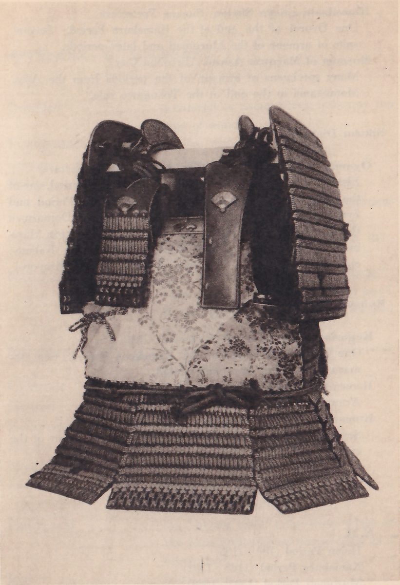 Black-and-white photograph of a suit of armour on a plain ground. A tall curved stack of dark laced bands stands at the right, and two upright dark plates, each with a small fan-shaped mark, stand at the center top. A pale body panel patterned with flowers and branches lies below them, and a skirt of laced bands with a woven lower edge flares out at the bottom.