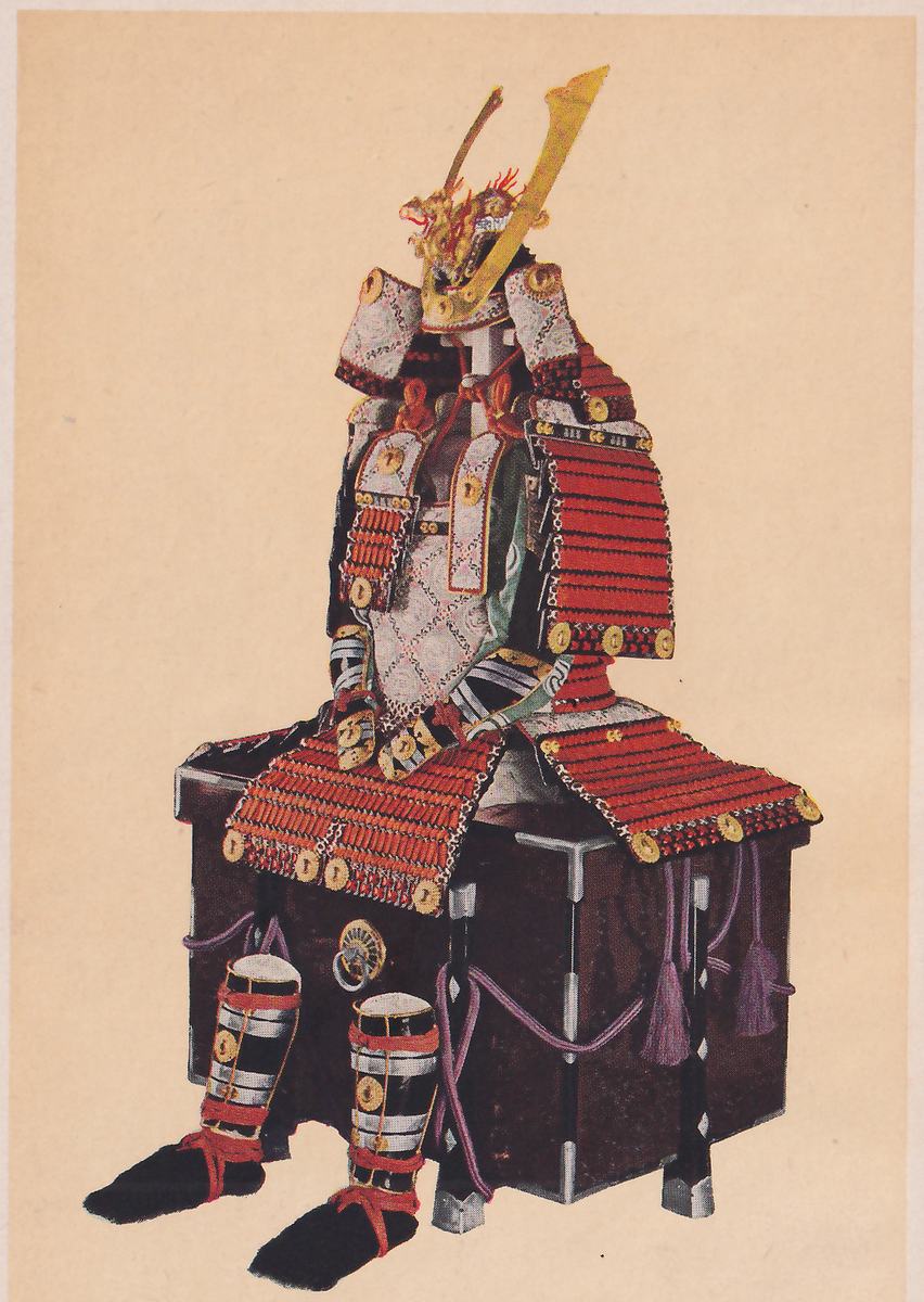 Color picture of a suit of armour set on a dark box with gray corner fittings and a round ring handle at its front, against a plain tan ground. A helmet at the top has a gold crest of curved prongs rising from its front. Rows of red-laced plates hang across the lap and at the sides, with pale panels patterned in small diamonds on the chest, and round gold discs along the plate edges. Purple cords with tassels hang down beside the box. In front of the box stand two black cylinders with pale bands and red ties, on dark flat pads.