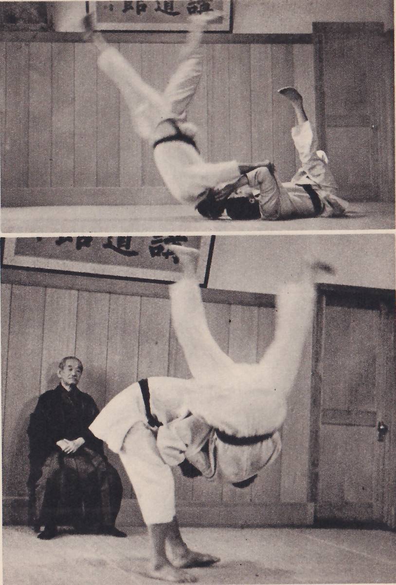 Two photographs, one above the other, in a hall with a framed Japanese sign on the wall. In the upper one a man in a judo suit is thrown over another's head, his legs flying up as the other man lies on the mat beneath him. In the lower one a man bent far forward carries his partner over his back, the partner upside down with his legs in the air, while an elderly man in a dark kimono sits watching at the left.