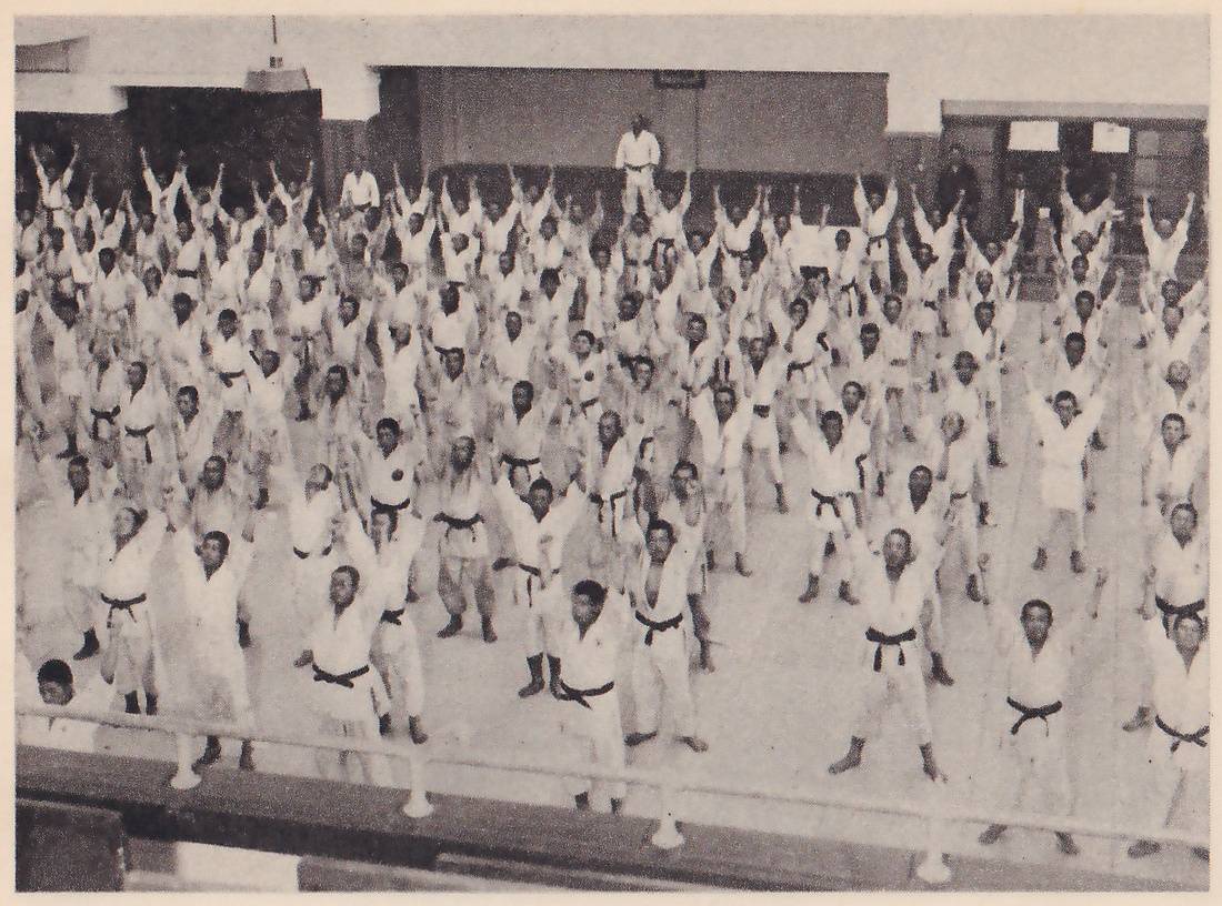 Seen from a gallery, rows of men in white judo jackets and dark belts stand across a hall floor, all with their arms raised overhead.