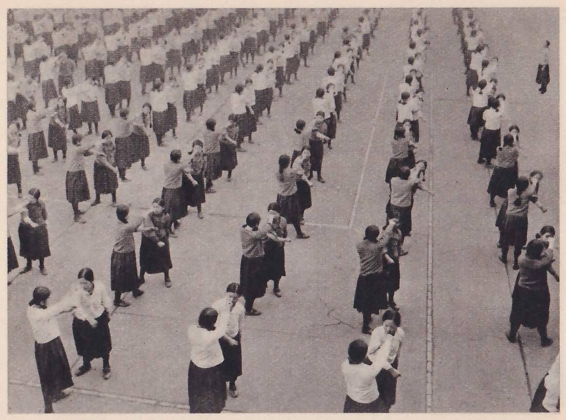 Seen from above, rows of schoolgirls in dark skirts and light tops stand on a paved yard in pairs facing each other, one arm out, in lines that run far into the distance.