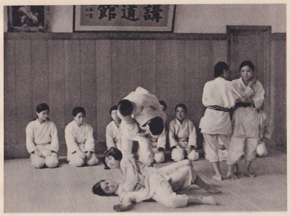 In a hall below a framed 講道館 sign, two girls in judo suits are in the middle of a throw, one on her back on the mat with the other bent over her; two more stand gripping each other at the right, and four girls kneel in a row along the wall.