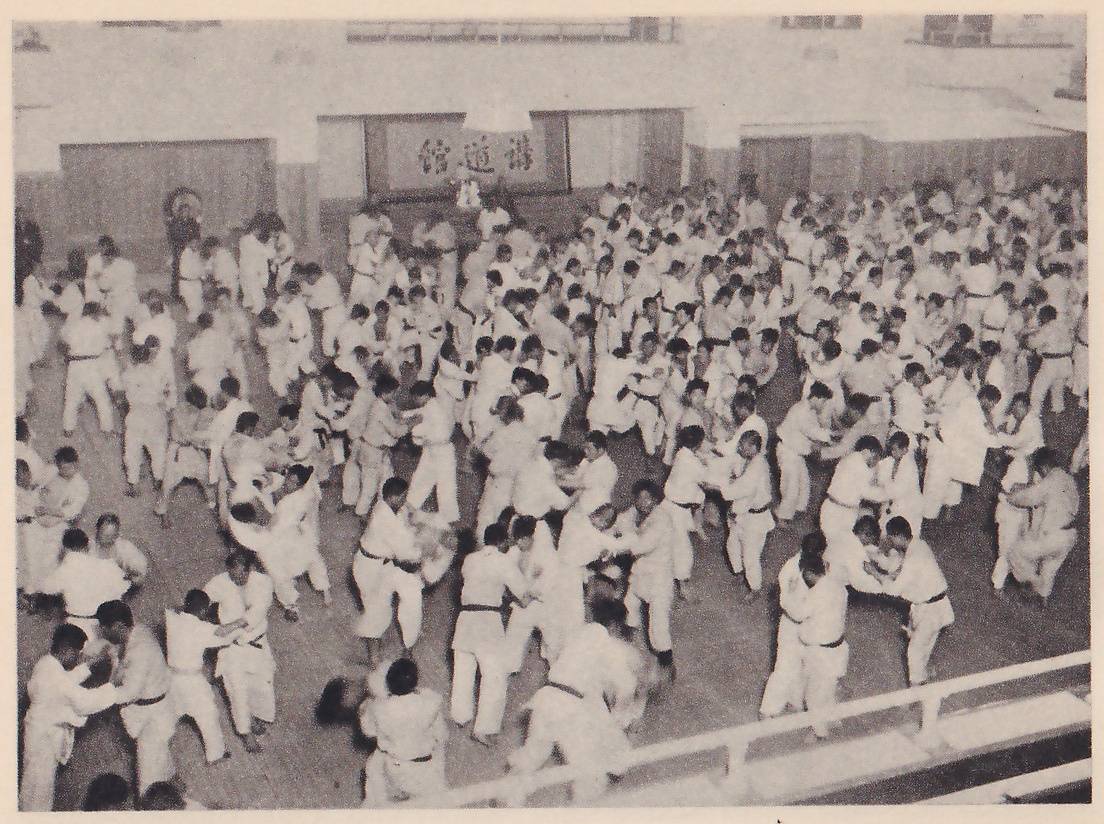 Seen from a gallery, hundreds of men in white judo suits practice throws in pairs across a large hall, with the 講道館 sign on the far wall.