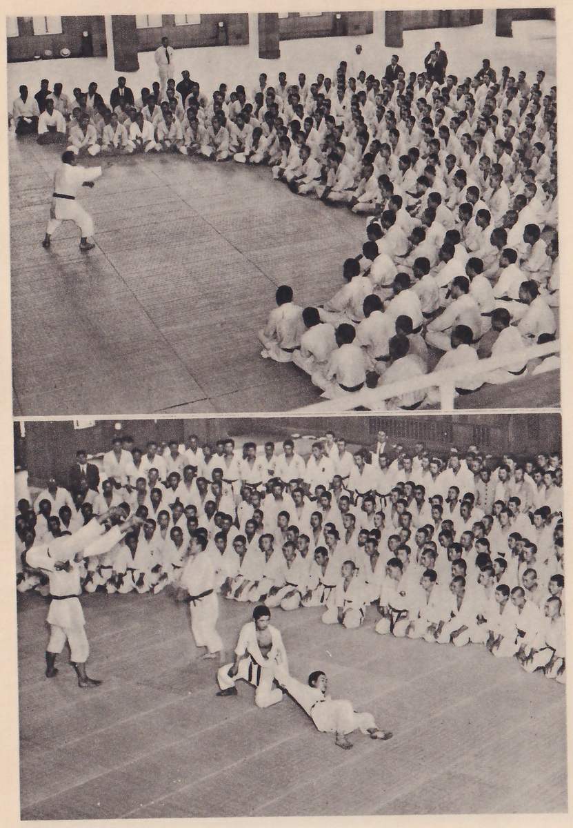 Two photographs, one above the other, of a large hall in which hundreds of men in white judo suits sit in a half circle on the mats. In the upper one a single man stands alone in the open space. In the lower one an instructor stands at the left with his arm raised while two others demonstrate a throw on the mat.
