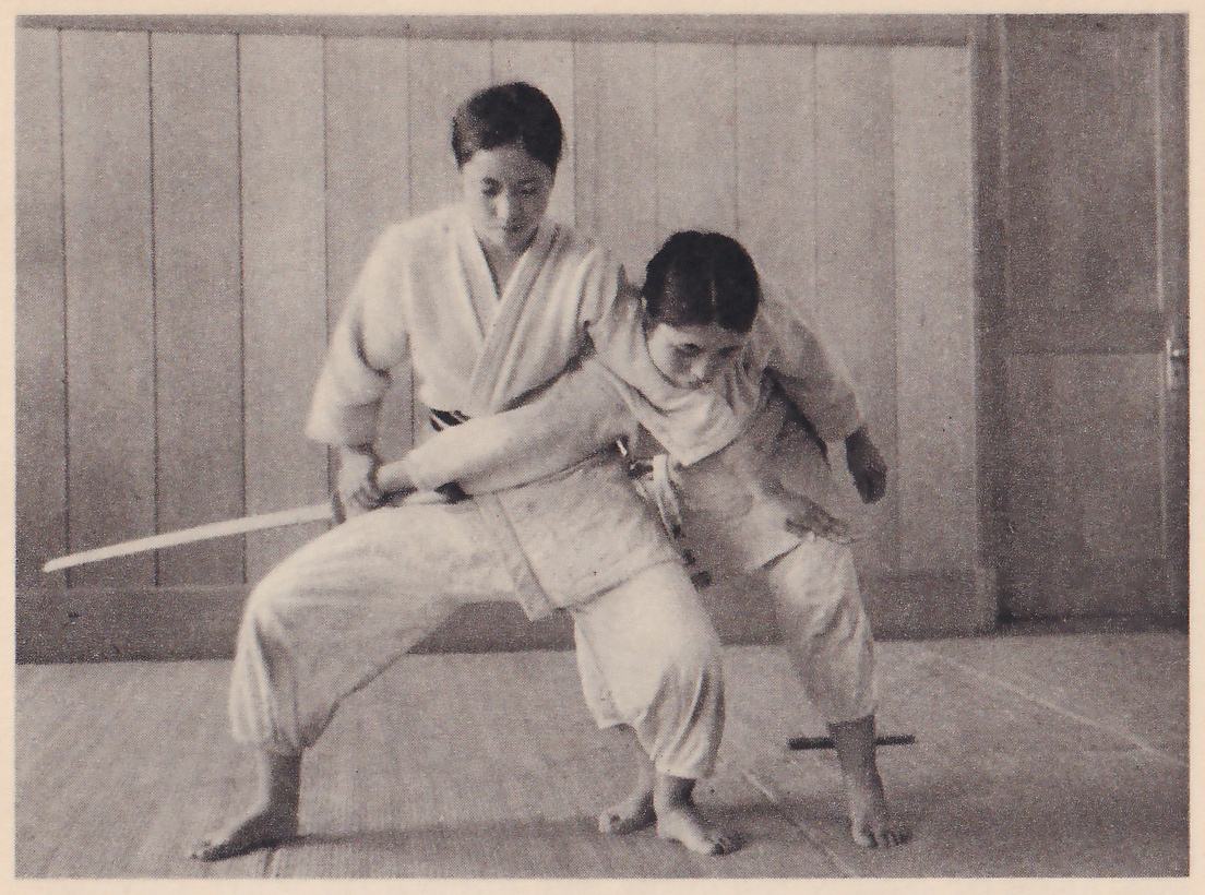 A woman in a judo suit stands with her knees bent wide, holding a wooden sword at her side with its blade out to the left, while a second woman bends forward against her and grips her sword arm at the wrist; a short wooden dagger lies on the mat behind them, and a wood-paneled wall is behind.