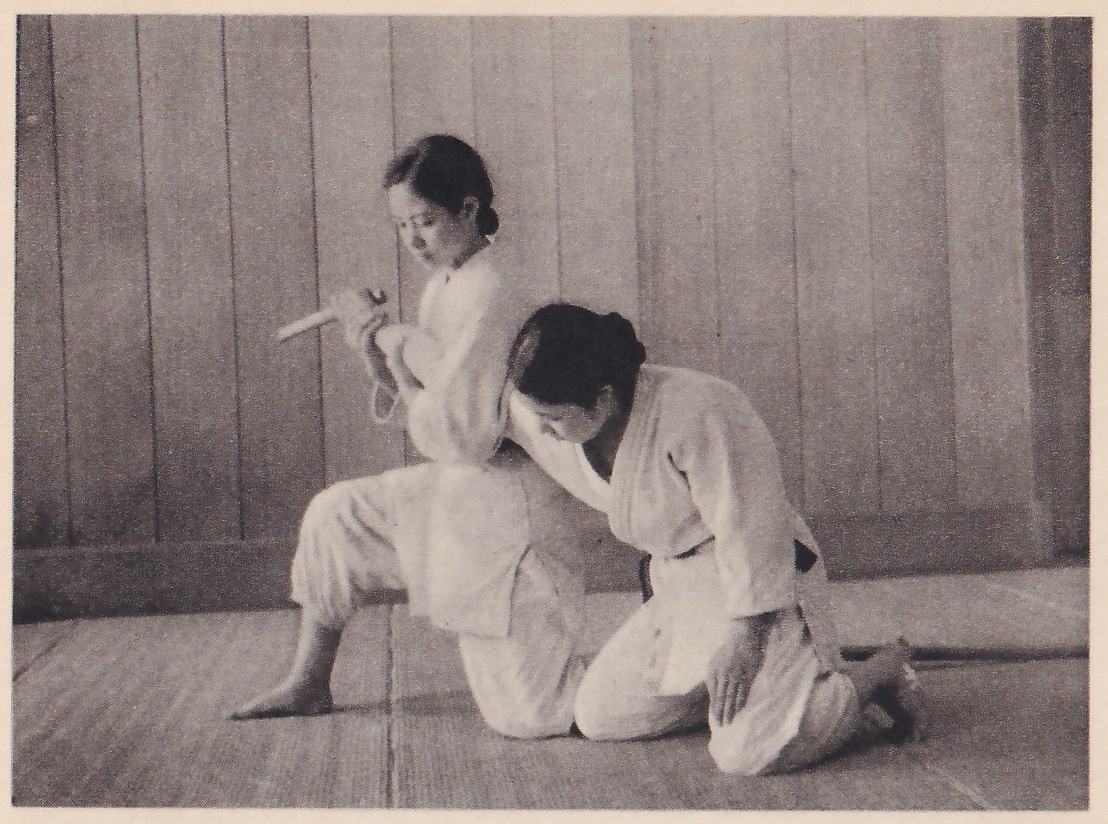 Two women in judo suits kneel on a mat. The one at the back holds a short wooden dagger raised at her side, while the one in front has pressed in against her and holds her arm.