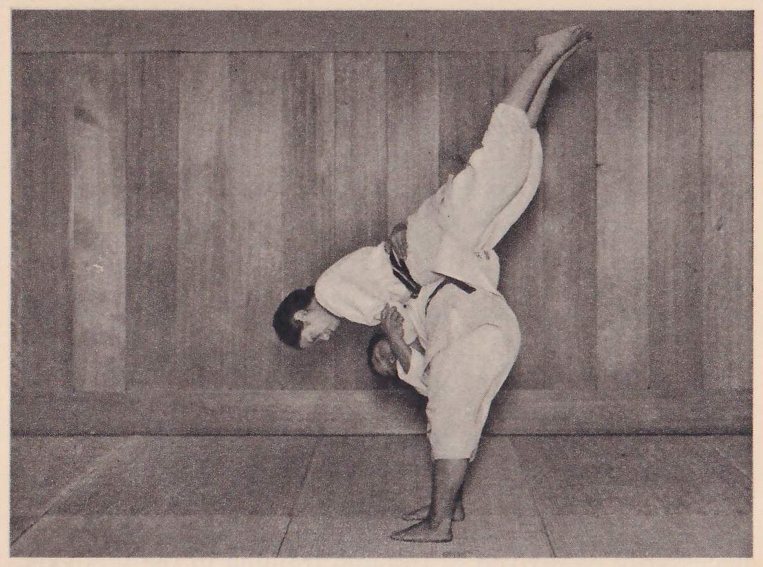 Against a wood-paneled wall, a person in a judo suit is thrown in mid-air, bent forward over the thrower's hip with one leg raised straight up.