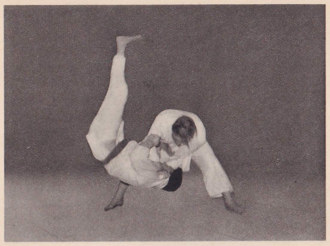 Against a plain dark background, one man in a judo suit bends forward and down while his opponent, in mid-air, has one leg raised straight up; the two are locked together at the middle.