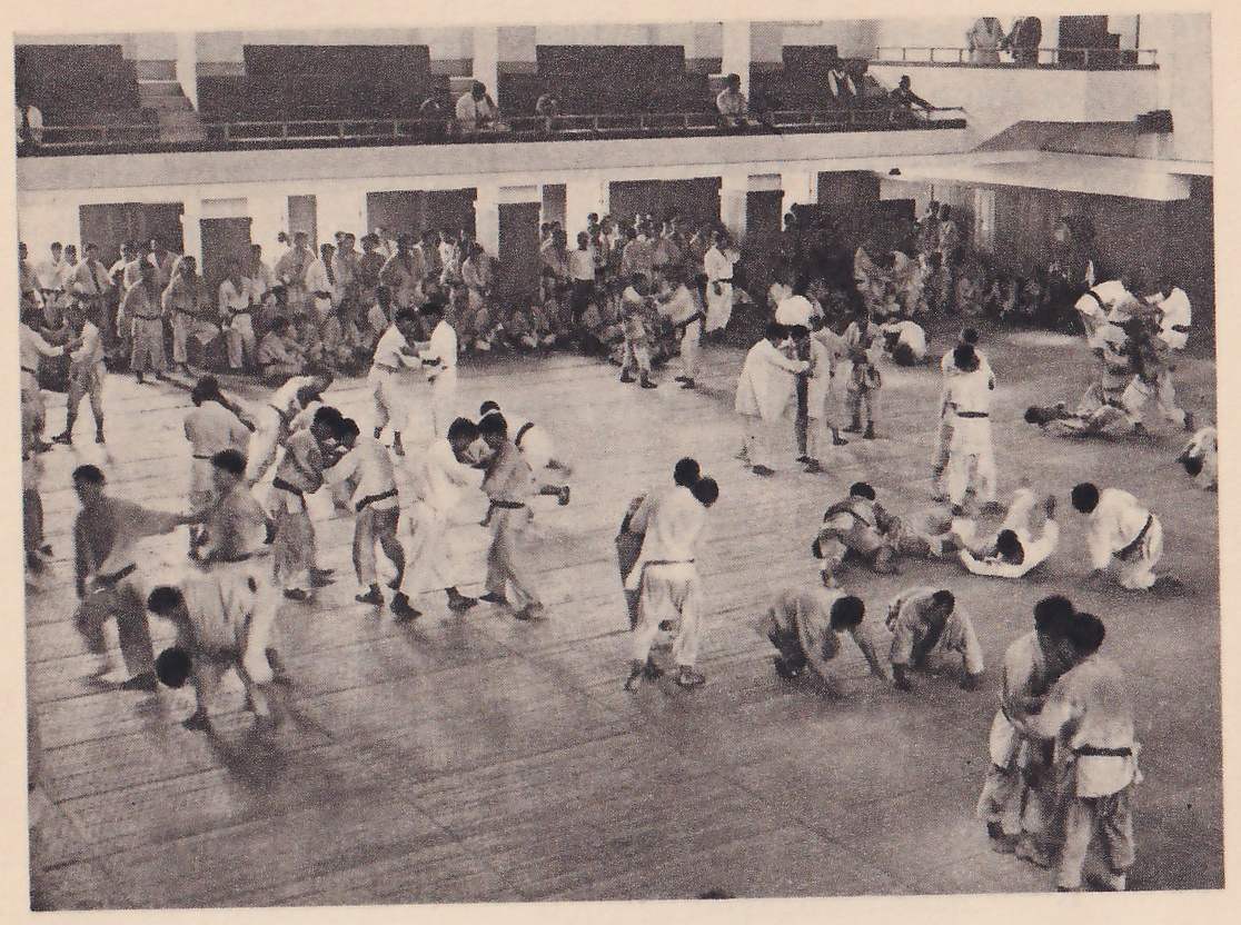 A large hall with galleries along the walls and a floor full of people in judo suits practicing in pairs, throwing and grappling, with spectators seated at the back.