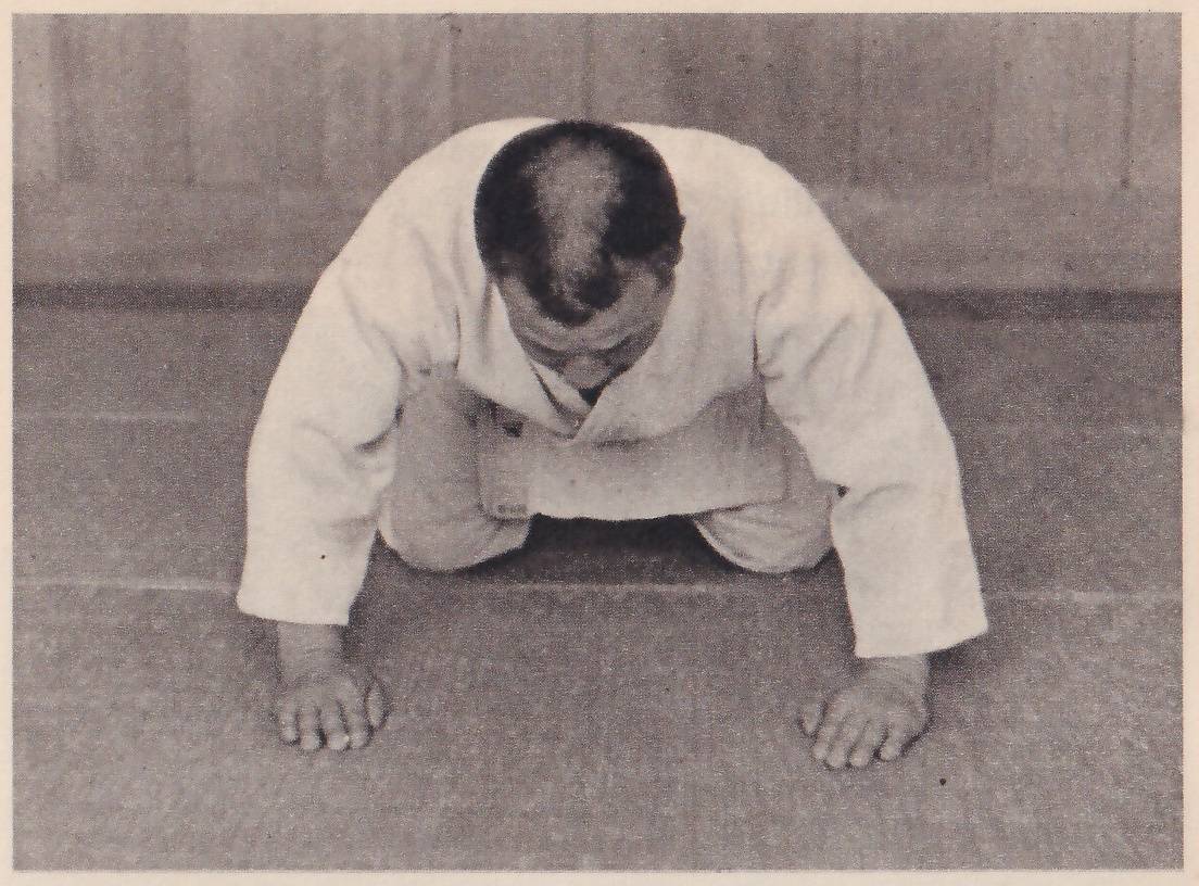 A man in a judo suit seen from the front kneels bent forward with his palms flat on the mat and his fingertips turned in toward each other, head down.