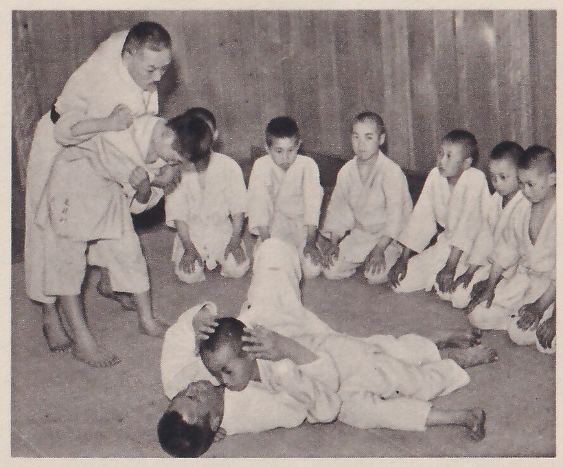 A man in a judo suit demonstrates a hold on a boy who stands bent against him; in the foreground one boy lies on his back on the mat while another boy leans over him and holds his head, and eight boys in judo suits kneel in a curve at the right, watching.