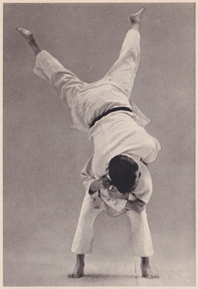 A man is thrown upside down in mid-air: his body is bent over the thrower's back and his legs are spread high in a V, while the thrower, bent forward with his feet apart, holds him against his own chest.