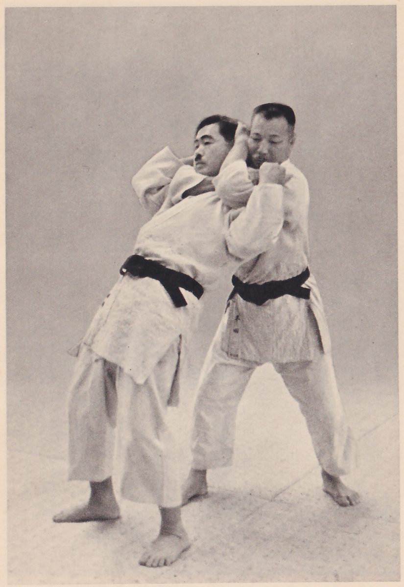 Two men in judo suits stand one behind the other. The man at the back has an arm around the other's neck from behind, so that the choked man's head is tilted back against him and his hand is raised to the arm.