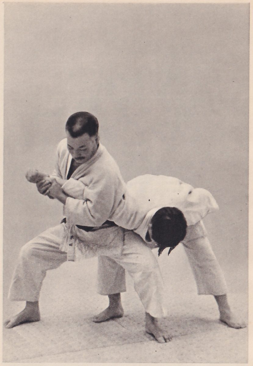 A man in a judo suit stands with his knees bent, gripping the arm of a second man, who is bent forward at the waist with his head down while his arm is held and twisted.