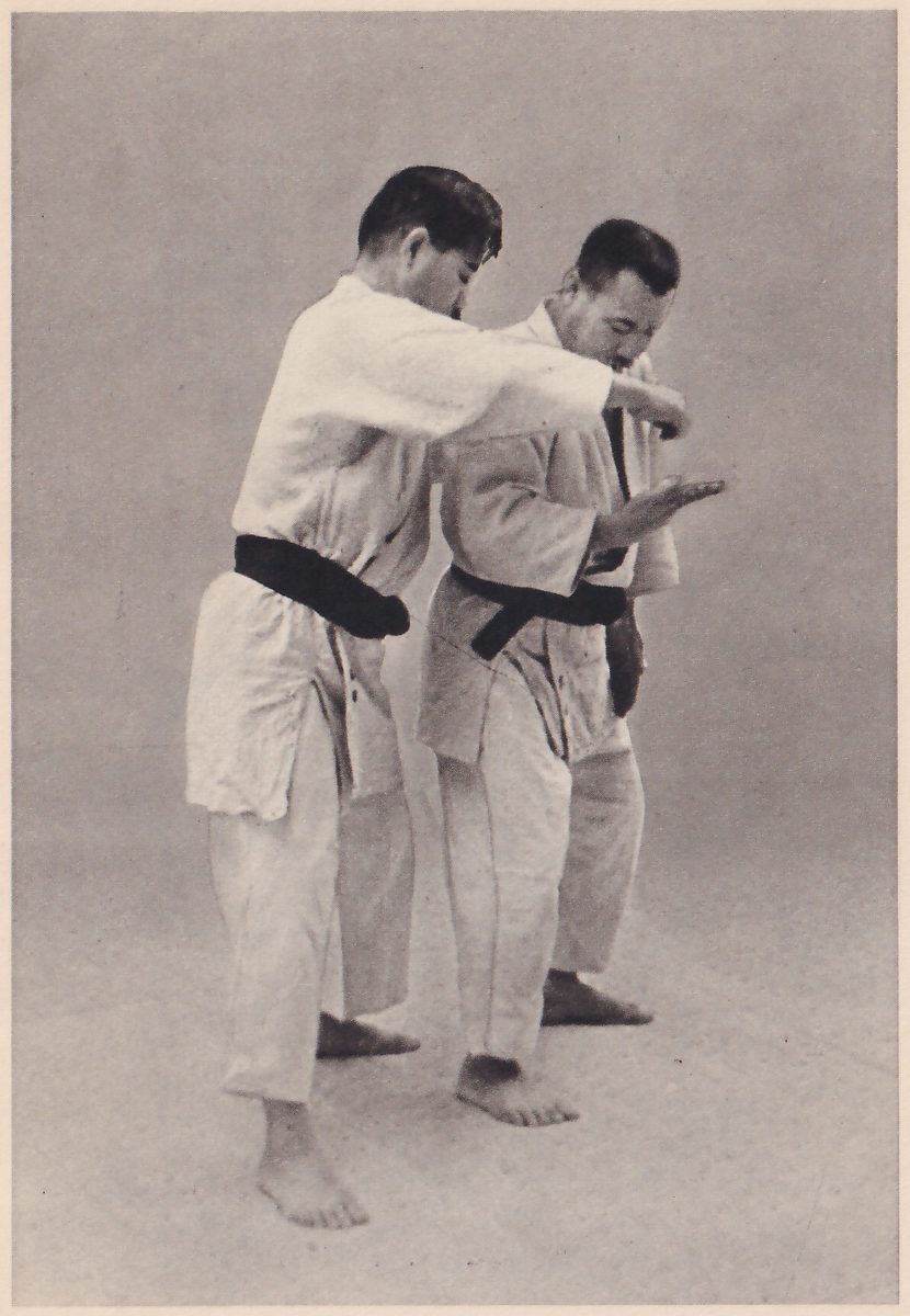 Two men in judo suits stand close together. One swings his bent arm toward the other, elbow forward, while the other holds up an open palm.