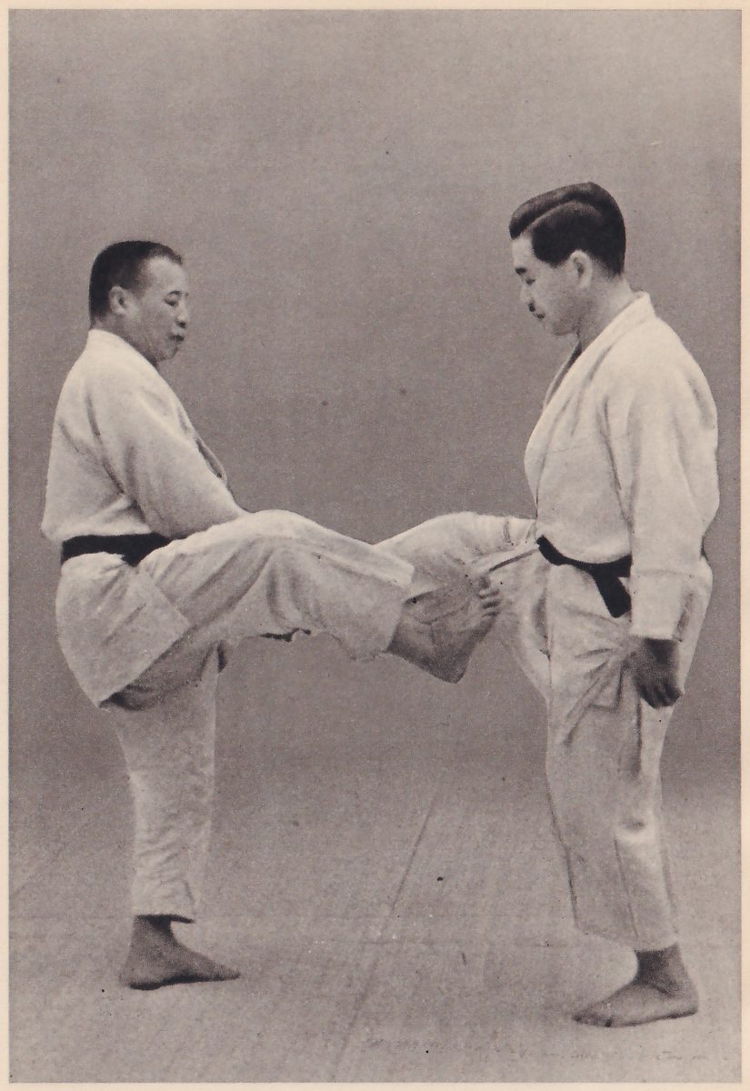 Two men in judo suits face each other. The man on the left stands on one leg and presses the ball of his other foot against the other man's lower belly, just below his belt; the other man stands with his hands at his sides.