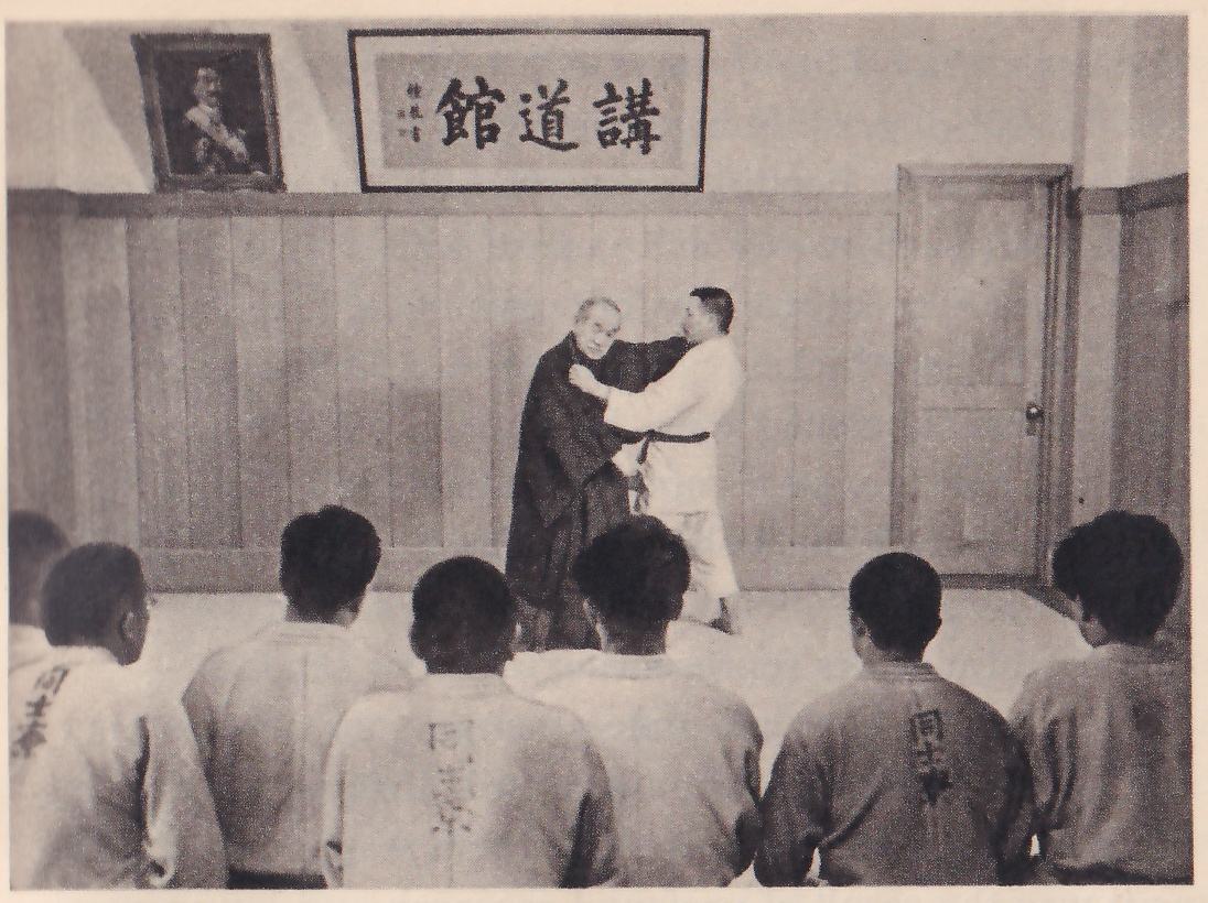 In a wood-paneled hall under a framed sign reading 講道館 (Kōdōkwan), an elderly man in a dark kimono holds the lapel of a standing man in a white judo suit and belt, while seven students in judo jackets sit on the floor with their backs to the camera, their jackets lettered on the back.