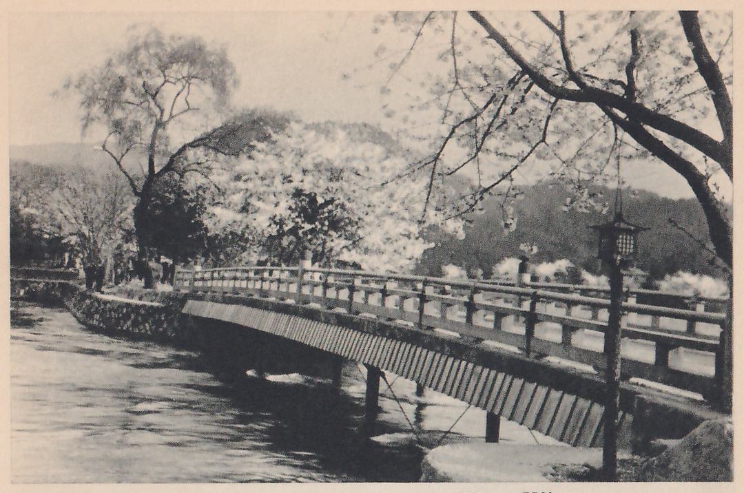 Black-and-white photograph of a bridge slanting from the center left to the lower right, with a railing along its length and a slatted side beneath, resting on short posts in rippling water. A hanging lantern with a lattice side stands on a thin post at the right, and a blossoming branch arches in from the upper right. Beyond the bridge, blossoming trees and tree-covered hills fill the background.