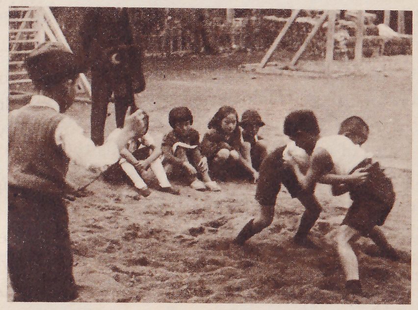 Photograph of bare ground in a playground. At the right two boys in shorts and light tops face each other, bent forward and gripping each other's arms. Four small children sit on the ground in a row in the center, one wearing a cap. At the left a man in a cap and a vest stands in the foreground with one arm raised toward them, and a dark-clothed figure stands behind. Climbing frames show at the top of the picture.