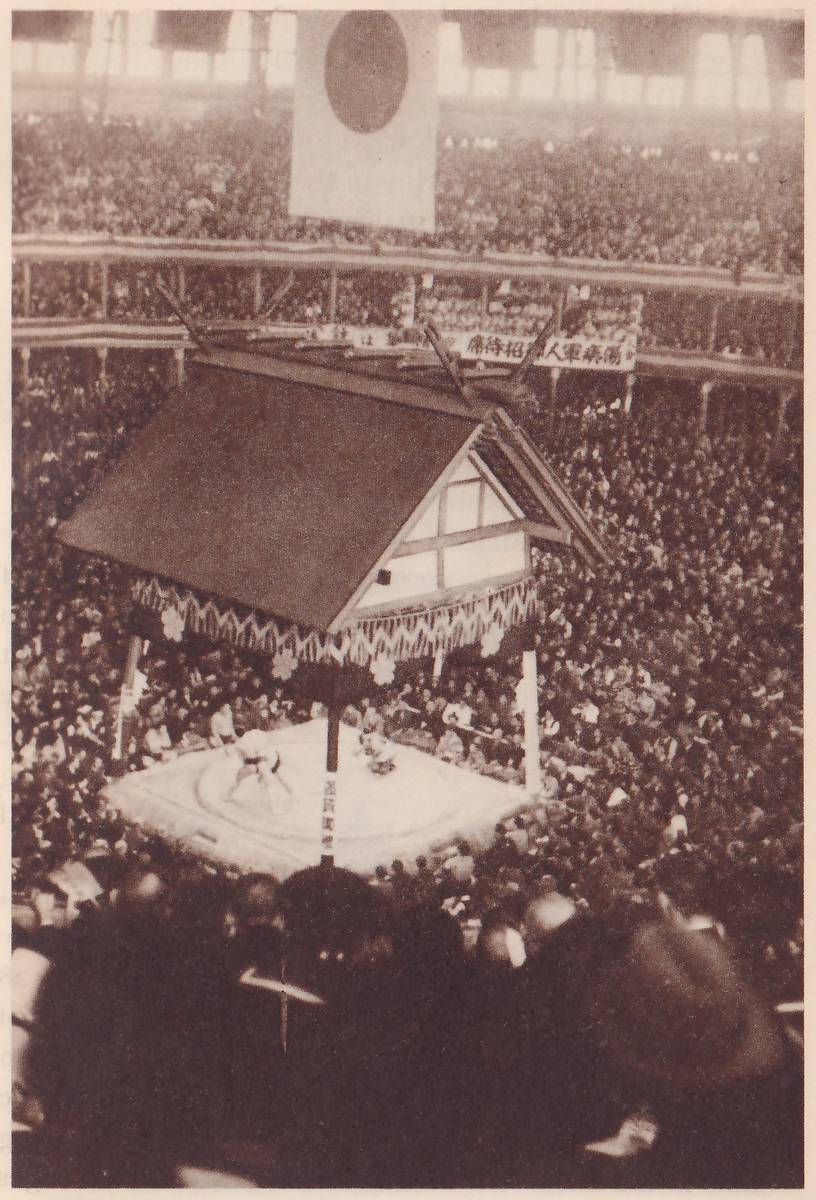 Photograph looking down into a very crowded hall. A square pale platform with two small figures on it stands at the center under a large roof with a patterned fringe along its eaves, with white flower-shaped marks on the fringe. A post with a vertical strip of writing stands at the front of the platform. A large flag with one dark disc hangs from the ceiling above, and a banner of Japanese characters runs along the gallery railing behind the roof. Spectators fill the galleries and the floor, and the dark heads and hats of the crowd fill the foreground.