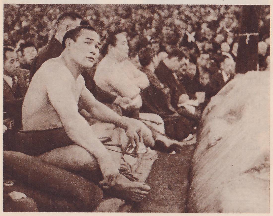 Photograph of a close crowd seen at floor level. In the left foreground a bare-chested man in a dark belt sits with his knees drawn up and his hands hanging in front of them, looking up and to the right. Behind him a large heavy man with bare shoulders sits with his arms folded, and behind and beside them men in dark suits and ties and in dark robes sit in a row. A large pale rounded surface fills the right edge, and a thick dark post stands at the upper right.