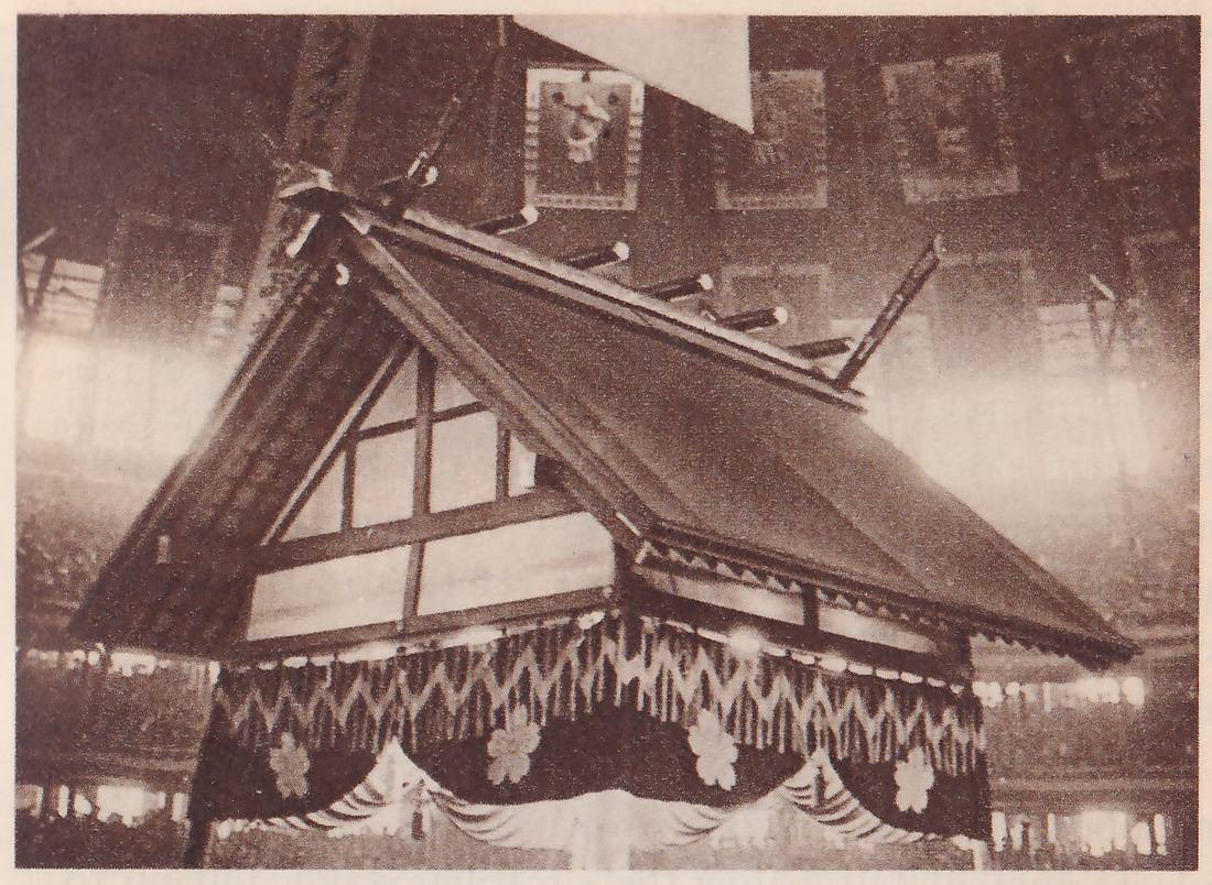 Photograph of a large gabled roof hanging in a hall, seen from below and a little to the side. Short round-ended beams stick out along its top ridge. Under the gable at the left, a wooden frame holds pale panels. A valance of zigzag-patterned cloth hangs from the eaves, with dark swags marked with white flower shapes under it. Behind the roof a dark wall carries several framed pictures, and a pale triangular flag hangs at the top. Hazy bright glare spreads across the right side.