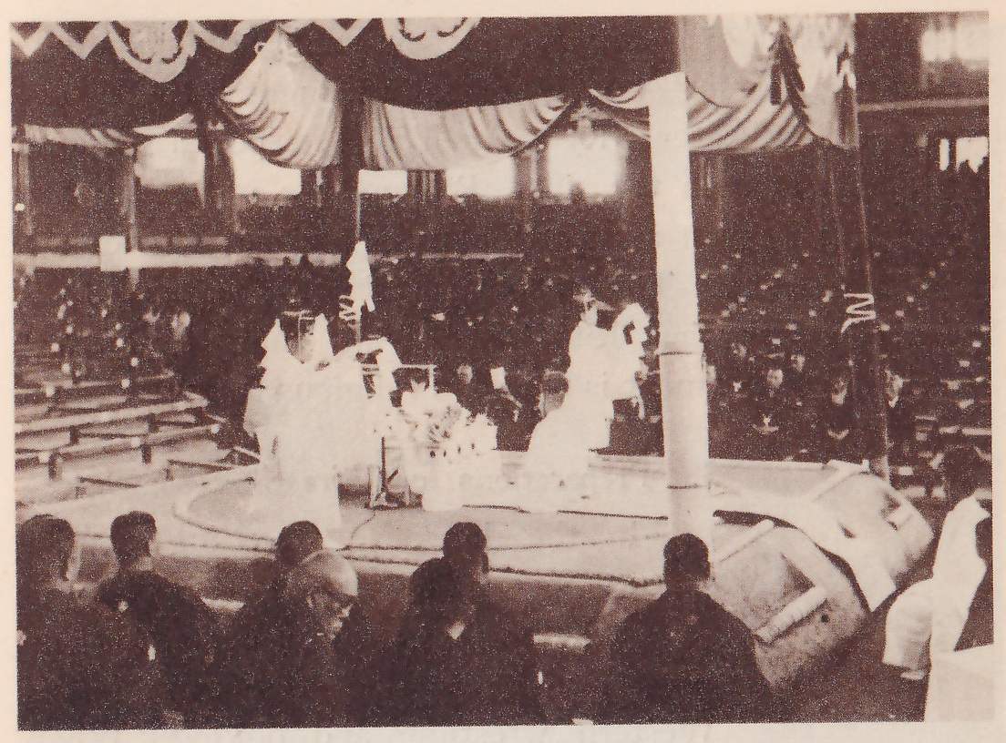 Photograph looking across a low platform from rows of seated people in dark clothes in the foreground. A thick pale column rises at the center right to a canopy of draped, gathered curtains with a zigzag edge across the top. On the platform several figures in pale robes stand or kneel, two at the left and one at the right of the column, with a small table between them holding pale shapes. Rows of benches and dark seated spectators fill the hall behind, and an irregular pale shape lies at the lower right.
