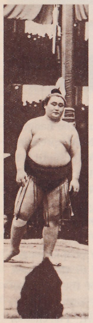 Tall narrow photograph of a heavy man standing facing the camera, bare to the waist, with a topknot. He wears a dark belt with a skirt of hanging strands from it, a wrapping around his left knee, and his arms hang at his sides. Behind him are a pale post and pieces of curtain with pale scalloped edges. A dark rounded shape fills the bottom edge of the picture below his feet.