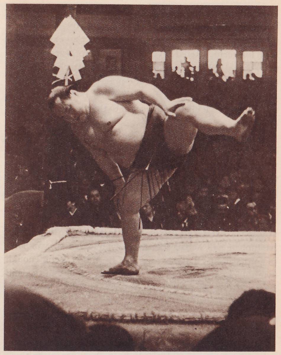 Photograph of a heavy, bare-chested man balancing on one foot on a raised flat platform. He is bent far forward with his head down at the left, his arm bent in front of him, and his other leg lifted out behind him to the right. A dark belt with hanging strands is at his waist. A bundle of folded white paper strips stands upright above his head. Behind him is a dark hall with lit windows and spectators, and dark heads fill the lower left and lower right corners.