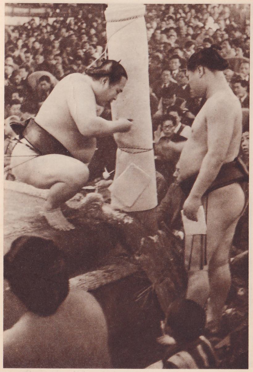 Photograph of two heavy bare-chested men in dark belts beside a thick pale post tied with rope. At the left one crouches on a raised surface with his arm stretched toward the post. At the right the other stands facing left, holding a folded sheet in one hand. Spectators fill the background, and dark heads and shoulders fill the lower left and bottom.