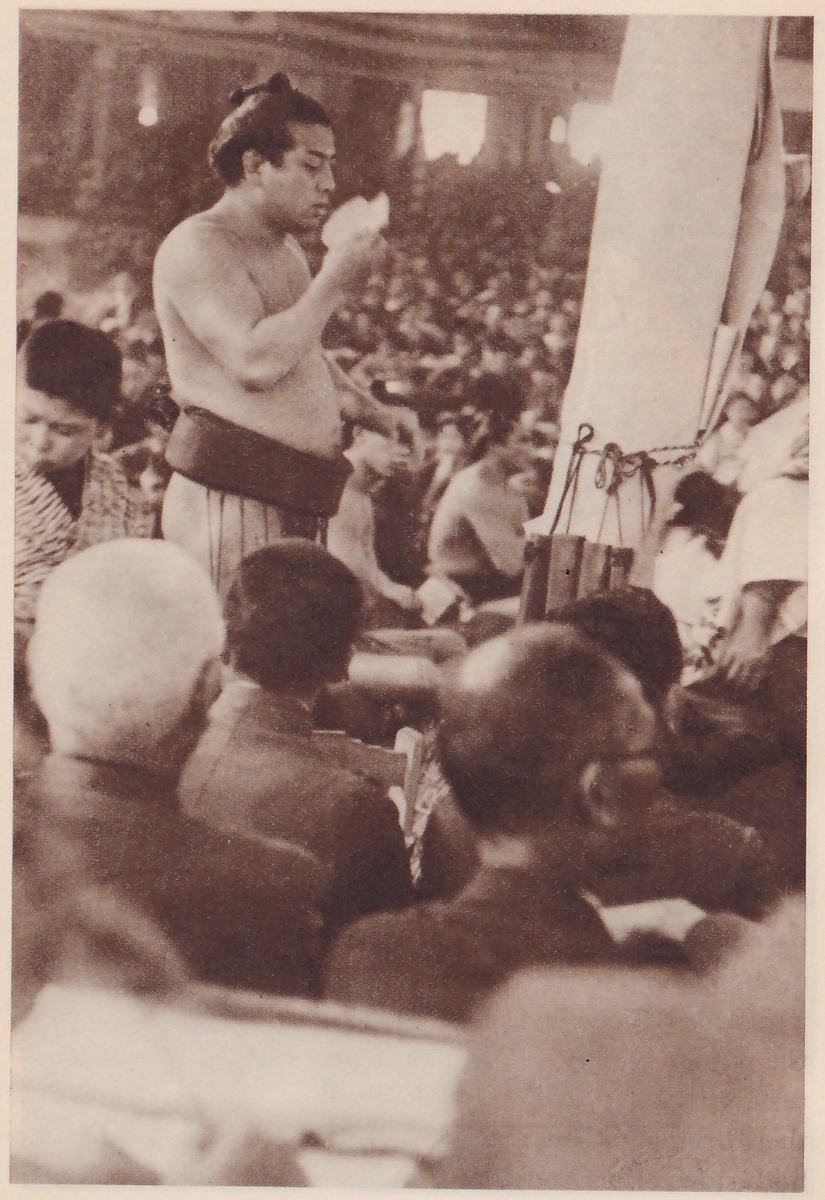 Photograph of a bare-chested man in a dark belt and striped trousers standing and holding a small pale sheet up to his mouth. A tall pale post tied with rope stands at the right, with a small wooden box at its foot. Behind him is a hall full of spectators. In the foreground several men in dark jackets are seen from behind, and a man in a striped robe is at the left edge.
