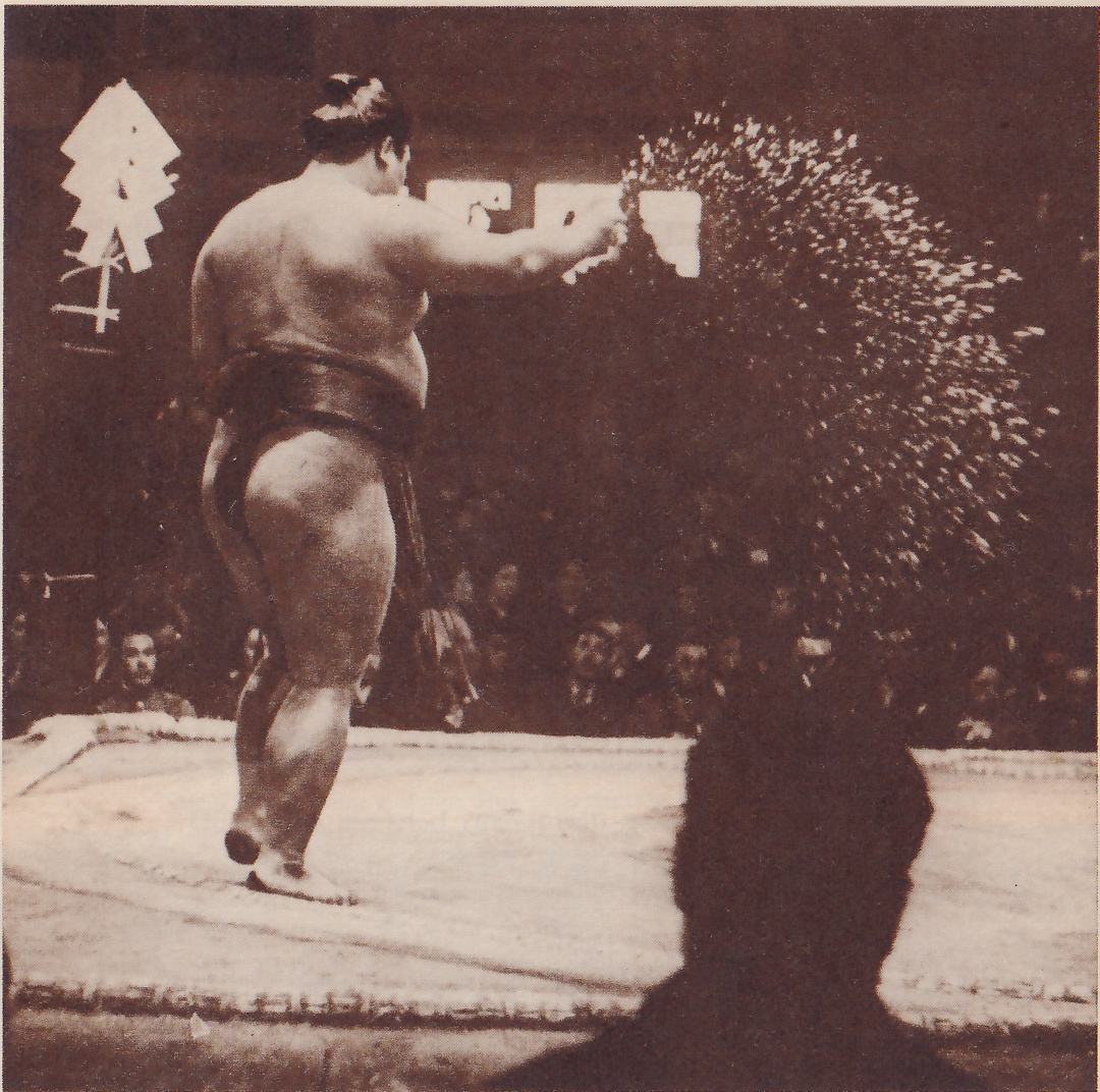 Photograph of a heavy man seen from behind on a raised flat platform, one arm stretched out to the right, with a wide arc of small white specks in the air beside his hand. His weight is on one foot, the heel of the other lifted. A dark belt with hanging strands is at his waist, and a topknot shows on his head. At the upper left a bundle of folded white paper strips stands on a small cross-shaped stand. Spectators sit in the dark behind, and the dark head and shoulders of a man fill the lower right foreground.