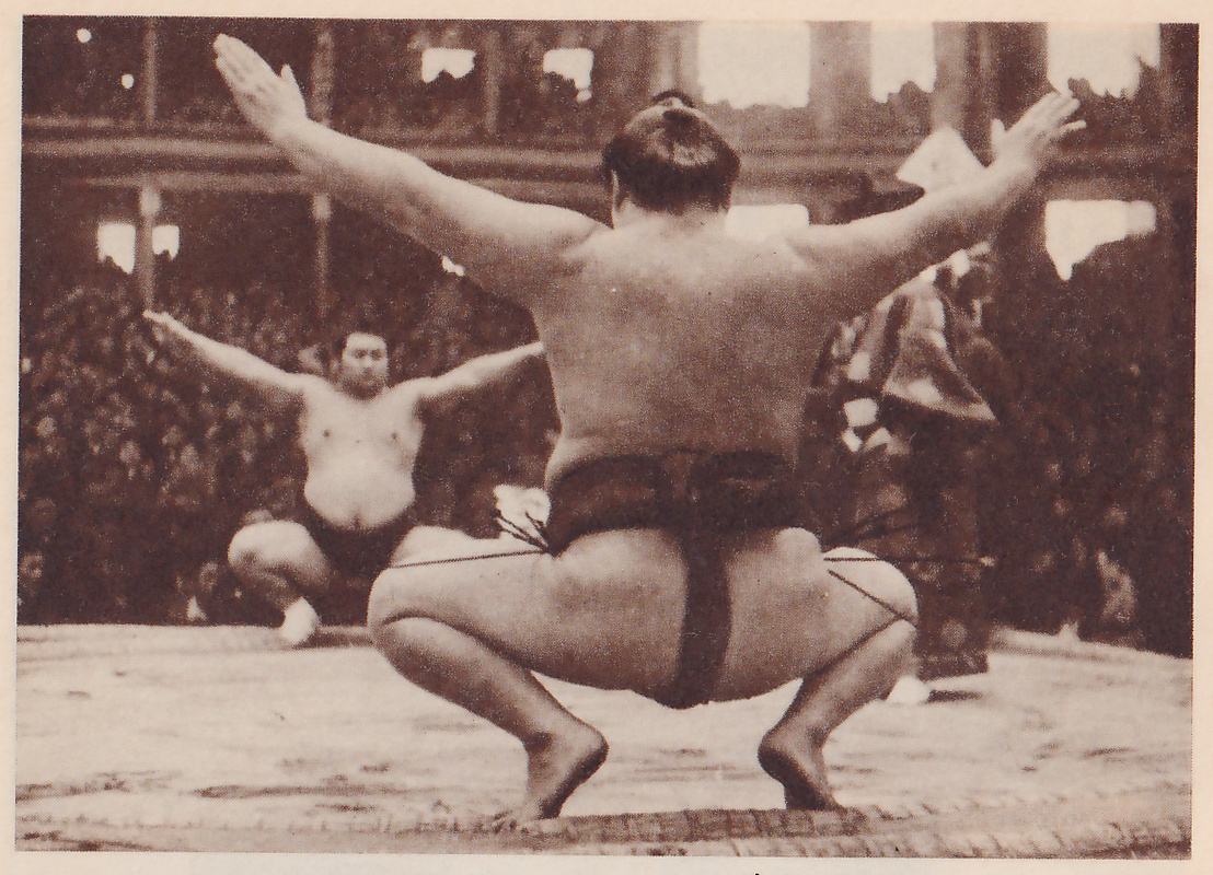 Photograph taken from behind a heavy man who squats low on a flat platform with his knees wide apart and both arms stretched out wide, palms open, his bare back to the camera and a dark belt at his waist. Farther away, a second heavy man squats facing him with his arms held out to the sides in the same way. A dense crowd fills the tiers behind them.