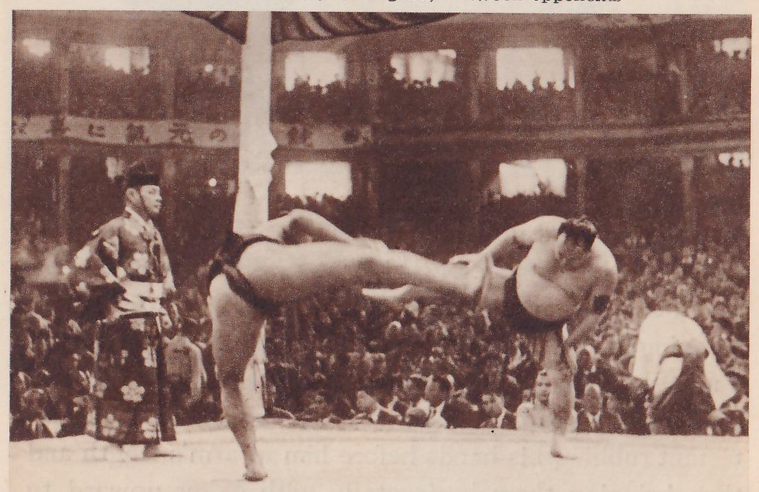 Photograph of two heavy bare men in dark belts at the center of a platform. The man at the left stands on one leg with the other leg lifted straight out to the right. The man at the right bends forward over his own planted leg, his arms bent in front of him. At the left a man in a long dark patterned robe and a black cap stands with his hands at his waist, and a pale column rises behind. A man in pale clothes bends low at the far right. Crowds fill the hall behind, and a long banner of Japanese characters runs along the gallery.