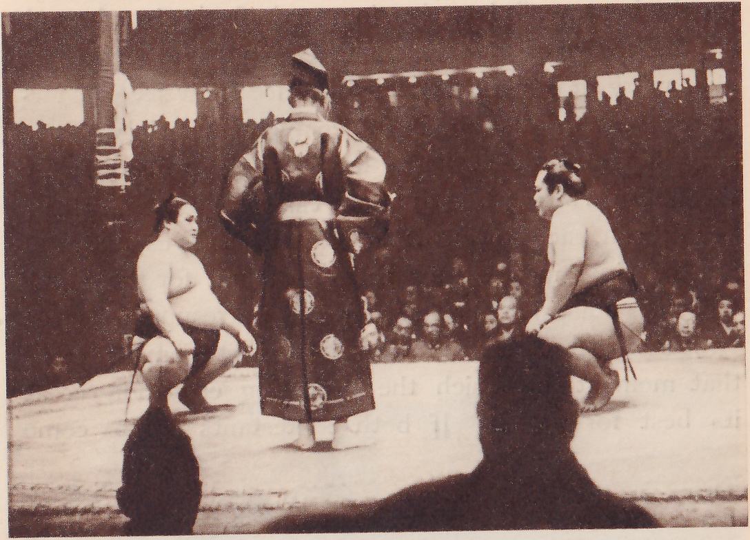 Photograph of two heavy, bare-chested men squatting on their heels at the left and right of a flat platform, each with his hands near his knees and his face toward the other. Between them a man seen from behind stands in a long dark robe marked with large round crests and a tall black cap, with his hands at his hips. A pale post stands at the upper left, spectators fill the background, and the dark heads of two men fill the lower foreground.