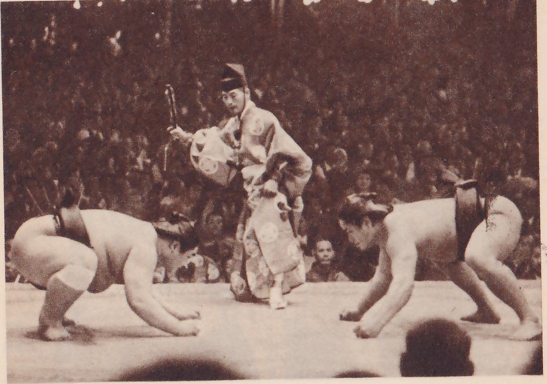 Photograph of two heavy men crouched facing each other on a platform, each leaning forward with his fists on the floor, the one at the left on both fists with his head level, the one at the right bent over lower. Between and behind them stands a man in a pale patterned robe and black cap, one hand holding a slim object raised at his side. A dense crowd fills the background, and the dark heads of two spectators show at the bottom.