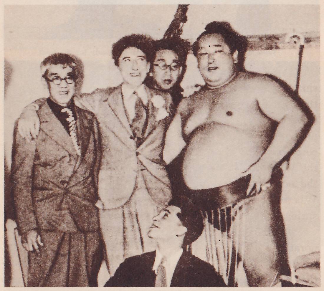 Photograph of five men posed close together. At the left stands a man with round dark glasses in a rumpled suit and a patterned tie. Beside him a man with curly hair, in a suit with a flower pinned to the lapel, has an arm over the first man's shoulder. Behind them a third man with glasses looks over their shoulders. At the right stands a very large, bare-chested man with his hand on his hip, wearing a dark belt with hanging strands. At the bottom center a man in a dark suit and tie sits low and looks up at the group. A pale wall is behind them.